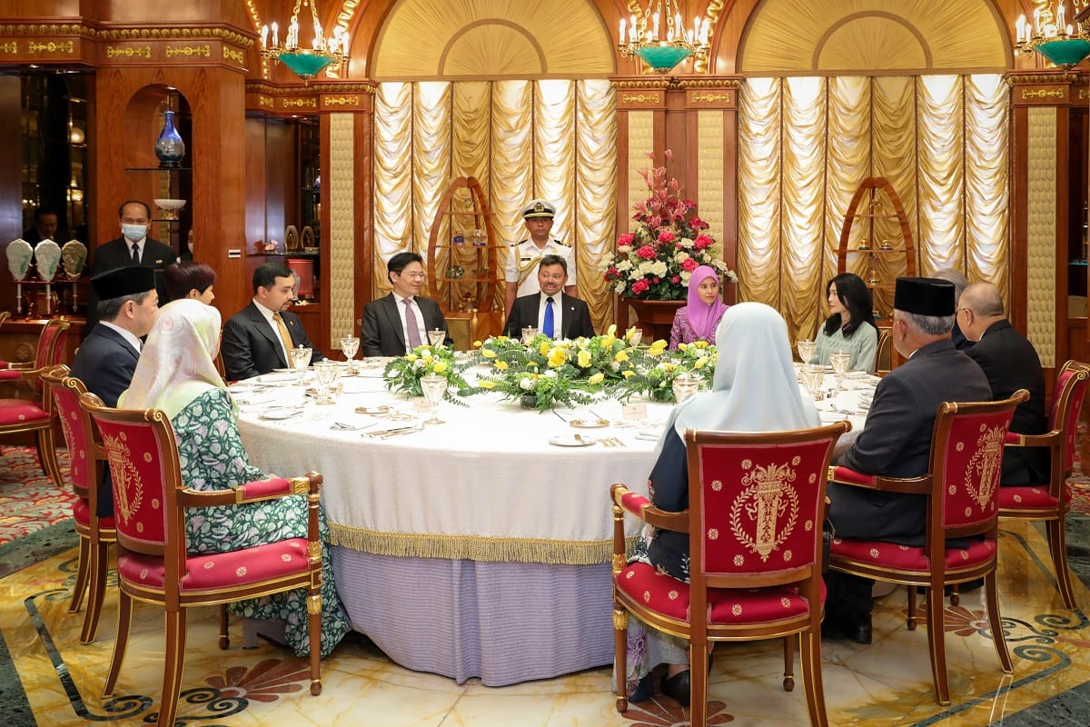 Dignitaries at round table with floral centerpiece in ornate room with uniformed service personnel.